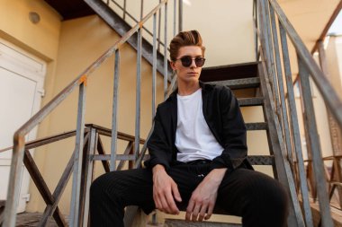 Demir merdivenlerde oturan zarif genç hipster adam. Bir yaz günü açık havada şık bir saç stili ile trendy güneş şık giysiler çekici adam modeli. Şık erkek giyim.