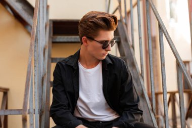 Şehrin demir bir merdiven üzerinde oturan beyaz bir T-shirt zarif bir siyah gömlek şık bir saç modeli ile trendy güneş gözlüğü genç yakışıklı hipster. Çekici adam. Şık erkek giyim.