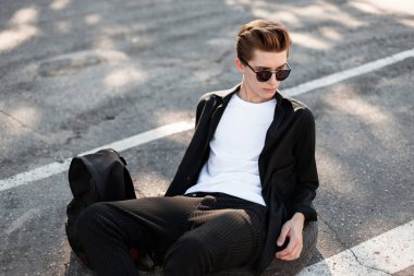 Trendy saç ile yakışıklı genç adam güneşli bir günde asfalt üzerinde rahatlatır. Bir sırt çantası ile şık kıyafetler moda hipster adam sokakta istirahat ve güneşin tadını çıkarın. Sokak modası.