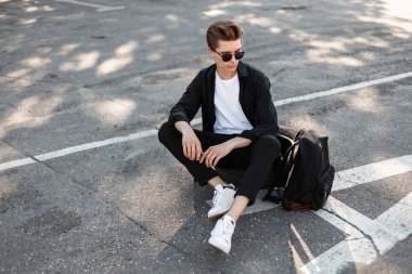 Güneşli bir gün şehirde Beyaz Ayakkabı dinlenme zarif moda giysiler içinde güneş gözlüğü saç modeli ile kentsel genç adam hipster. Güzel adam güneşte asfaltta oturan rahatlatır. Sokak stili.