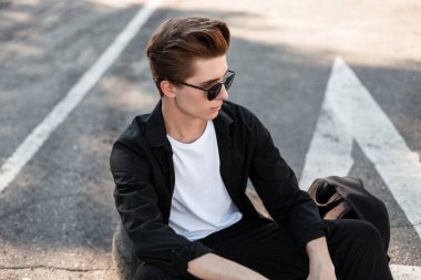 Koyu güneş gözlüğü siyah bir gömlek şık bir saç modeli ile çekici genç adam şehirde sokakta asfalt oturan dinleniyor. Yakışıklı Avrupa adam yaz güneşi açık havada sahiptir. Stil.