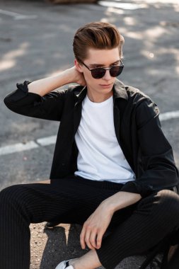 Güneşli bir yaz gününde bir şehirde asfalt üzerinde oturan bir saç modeli ile trendy güneş gözlüğü zarif giysiler genç yakışıklı şık adam. Modern kentsel adam hipster sokakta dinleniyor. Sokak stili