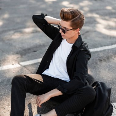 Saç modeli ile sırt çantası ile pantolon trendy gömlek genç hipster adam parlak bir yaz güneşli bir gün şehirde asfalt oturan pozlar. Güneş gözlükleri yakışıklı hipster adam güneşte rahatlatır.