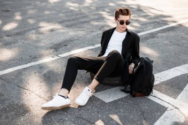 Güneş gözlüğü bir sırt çantası ile beyaz trendy spor ayakkabıları zarif siyah kıyafetler modern genç adam hipster sokakta bir lastik tekerlek üzerinde oturur. Kentsel adam bir yaz gününde açık havada güneşin içinde rahatlatır.