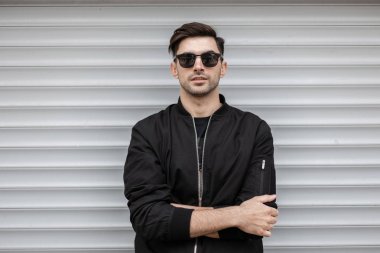 Moda bir siyah ceket koyu güneş gözlüğü şık bir saç modeli ile Amerikan Genç esmer adam şehirde beyaz metalik duvar yakınında duruyor. Çekici modern hipster adam. Trendy erkek giyim.