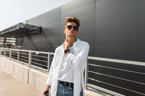 Europese stijlvolle jonge hipster man in modieuze zomer kleren in zwarte zonnebrillen met een modieuze kapsel staat op de straat op een zonnige dag. Aantrekkelijk Guy fashion model. Jeugd Streetstyle. — Stockfoto