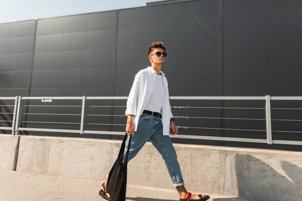 Jonge stijlvolle man hipster in trendy kleding in zonnebril met een zwarte doek zak in Vintage sandalen wandelingen is in een stad in de buurt van een grijs gebouw. Amerikaanse kerel. Modieuze zomer herenkleding. Streetstyle. — Stockfoto