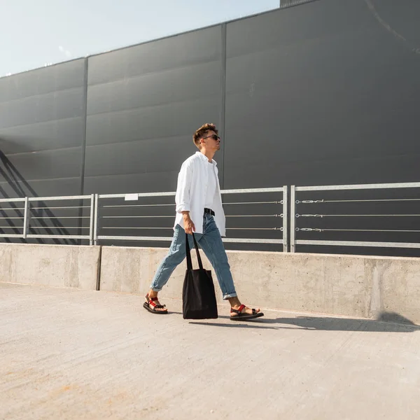 Joven hipster de moda en gafas de sol en ropa de verano de moda con bolsa de tela en sandalias vintage camina por la calle en un día soleado. Un tipo moderno. Nueva colección de ropa de hombre con estilo de verano . — Foto de Stock