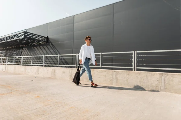 Νέος Ευρωπαίος άνθρωπος μοντέλο μόδας σε μοντέρνα τζιν σε ένα κομψό πουκάμισο σε μοντέρνα σανδάλια σε γυαλιά ηλίου με τσάντα βόλτες κοντά σε ένα γκρίζο κτίριο. Ένας τύπος από το αστικό. Νέα καλοκαιρινή συλλογή από αντρικά ρούχα — Φωτογραφία Αρχείου