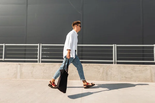 Elegante joven hipster en ropa de moda en sandalias de cuero con una bolsa de tela en gafas de sol camina por la calle cerca de un edificio gris. Hombre urbano de moda viaja por la ciudad en un día soleado de verano — Foto de Stock