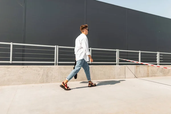 Jonge man met een trendy kapsel in een wit overhemd in Vintage blauwe jeans in lederen sandalen met een zwarte tas loopt langs de straat in de buurt van een grijs gebouw. Moderne stijlvolle kerel op een wandeling op een zonnige dag. — Stockfoto