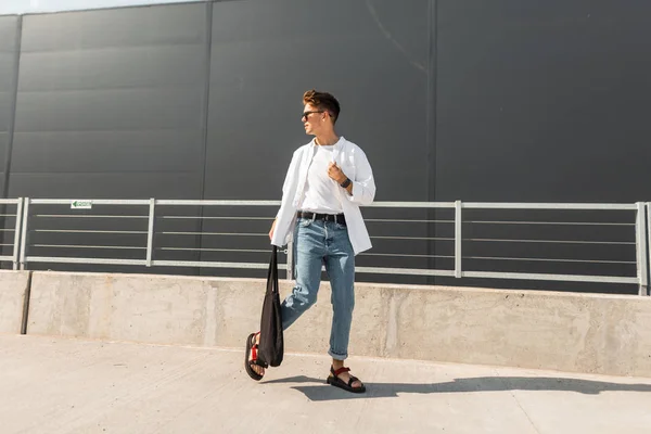 Νέοι μοντέρνος άνθρωπος μοντέλο σε ένα λευκό vintage shirt σε μπλε κομψό τζιν σε γυαλιά ηλίου με μια μαύρη τσάντα περπατάει στο δρόμο σε μια ηλιόλουστη μέρα. Ωραίος χίπστερ. Μοδάτο καλοκαιρινό βλέμμα. Μόδα της νεολαίας. — Φωτογραφία Αρχείου