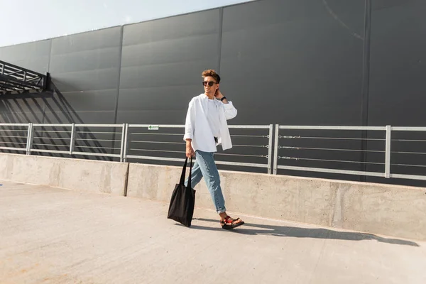 Joven joven de moda camina por la ciudad en un día soleado. Guapo chico hipster en ropa de moda en zapatos de verano con una bolsa en gafas de sol está viajando cerca de un edificio gris. Verano juventud mirada . — Foto de Stock