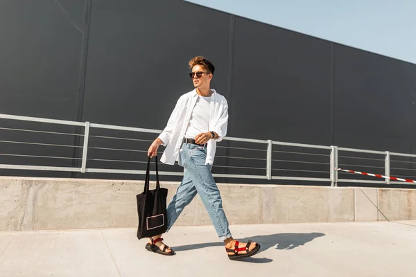 Moderne jonge man in stijlvolle kleren in zonnebrillen in rood lederen sandalen met een vintage zwarte tas reist op een heldere zonnige dag in de buurt van een grijs gebouw. Trendy hipster Guy wandelingen. Modieuze herenkleding. — Stockfoto