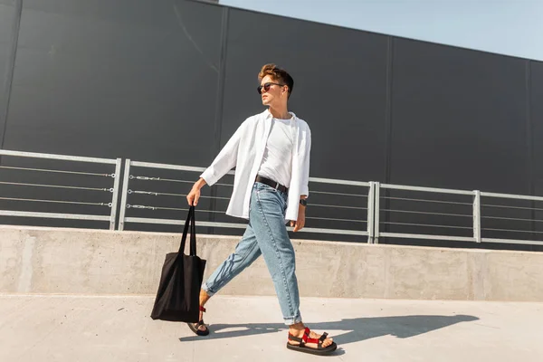 Americký módní mladík ve stylových šatech v červených sandálech s látkový vak v slunečních brýlích cestuje na ulici poblíž šedé budovy. Stylový chlapík venku. Nová sbírka letních oděvů. — Stock fotografie