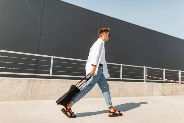 Joven hombre hipster europeo en una camisa blanca en vaqueros azules en sandalias de moda con una bolsa negra camina por la calle en un día soleado. El tipo atractivo disfruta de un paseo por la ciudad. Moda masculina de moda . — Foto de Stock