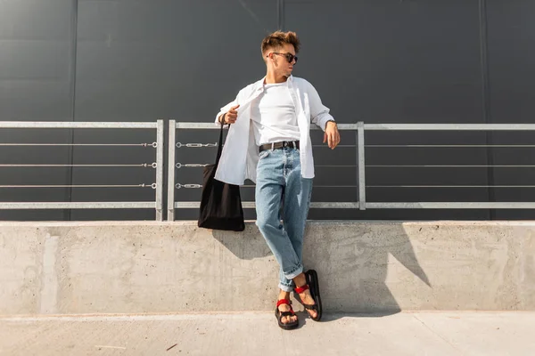 Europese Young Man model in stijlvolle kleding in lederen rode sandalen met een zwarte tas in zonnebril rust in de buurt van de metalen reling in de stad. Knappe hipster vent. Modieuze zomer herenkleding. — Stockfoto