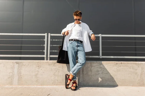 Joven hipster moderno en ropa blanca de verano de moda en gafas de sol con elegantes sandalias de cuero rojo con una bolsa se encuentra en un día soleado cerca de un edificio gris. Buen tipo. Moda masculina de moda. Estilo de calle — Foto de Stock