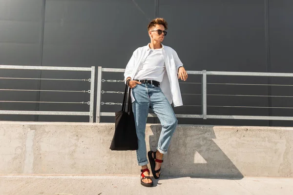 Joven urbano hipster con un peinado en ropa de moda con una bolsa de pie cerca de la barandilla de metal cerca del edificio gris. Un tipo moderno de moda. Moda europea juvenil . — Foto de Stock