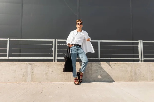 Modieuze stedelijke jonge man in rode sandalen in een wit overhemd in blauwe jeans in zonnebrillen met een zwarte tas ontspant in de straat op een heldere zonnige dag. Stijlvolle hipster vent. Zomerlook. Jeugd stijl. — Stockfoto