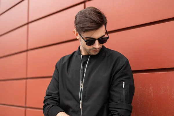 Der junge, gut aussehende Hipster mit stylischer Frisur steht in einer schwarzen, trendigen Jacke mit dunkler Sonnenbrille auf der Straße in der Nähe des roten modernen Gebäudes. Stadtmensch spaziert durch die Stadt. — Stockfoto