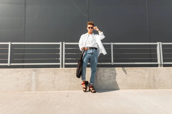 Jonge moderne hipster man in een wit overhemd in blauwe jeans in zonnebrillen met een zwarte tas in rode sandalen staat op een zonnige dag in de stad. Knappe kerel model. Trendy zomerlook. Jeugd mode. — Stockfoto
