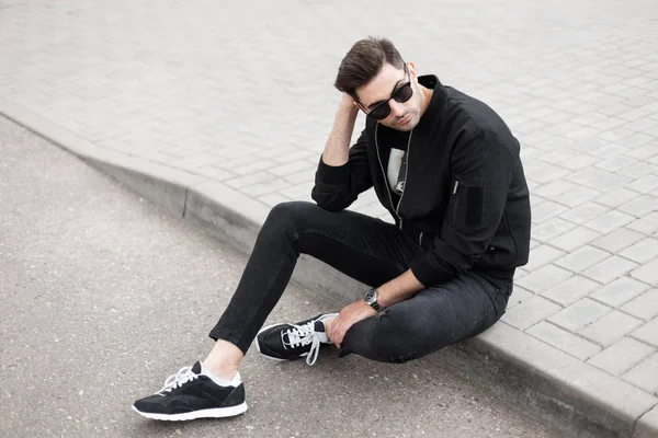 Cool guapo joven hipster hombre en una chaqueta elegante en vaqueros negros en zapatillas de deporte con gafas de sol de moda está sentado en la acera en una calle en el día de verano. Buen chico modelo es relajante al aire libre . — Foto de Stock