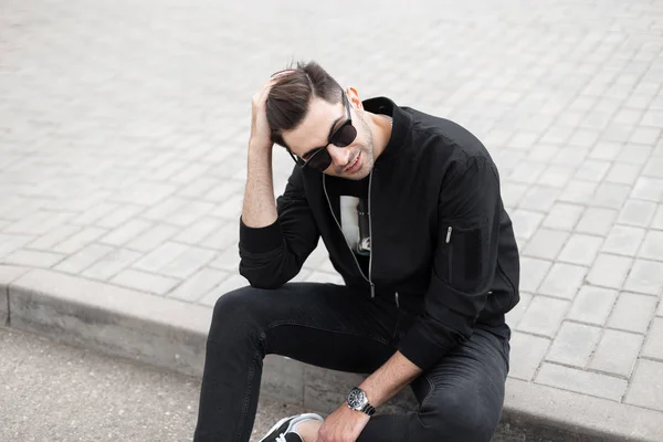 Przystojny młody człowiek Hipster z modną fryzurę w modnej czarnej kurtki w stylowych okularów przeciwsłonecznych siedzi w płytkach i prostuje włosy. Miejski facet w stylowe ubrania spoczywa w mieście. Stylu. — Zdjęcie stockowe
