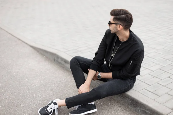 Stylischer junger Hipster in schwarzer Jacke, schwarzer Jeans und Turnschuhen mit trendiger Sonnenbrille sitzt an einem Sommertag auf dem Bürgersteig einer Straße. Stadtmodell entspannt sich im Freien. — Stockfoto