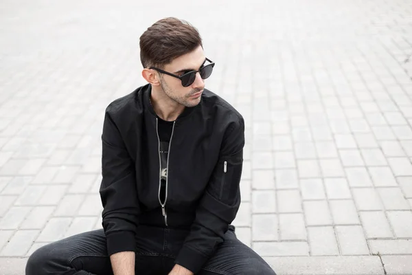 Modern cool young hipster man in stylish sunglasses in black fashionable jacket in jeans is resting sitting on a tile in a city on a bright summer day. Trendy american model guy relaxes outdoors. — Stock Photo, Image