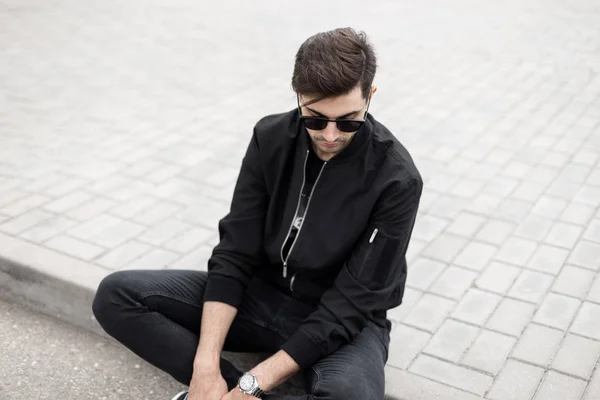 Attraktiva unga hipster man i snygga solglasögon i svart fashionabla jacka i jeans vilar sitter på en bricka på en gata på en solig sommardag. Trendig amerikansk modell kille slappnar utomhus. — Stockfoto