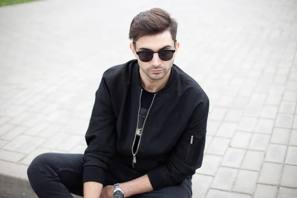 Young model man in fashionable black clothes in dark sunglasses rest sitting on a tile in the city on a bright summer day. Handsome American guy model enjoys the weekend outdoors. Trendy menswear.