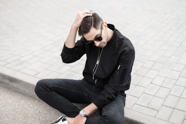 American Young Man model met een stijlvolle kapsel in een modieuze zwarte jas in trendy zonnebrillen zit in een tegel en recht haar. Knappe hipster kerel in stijlvolle kleren rust in de stad — Stockfoto