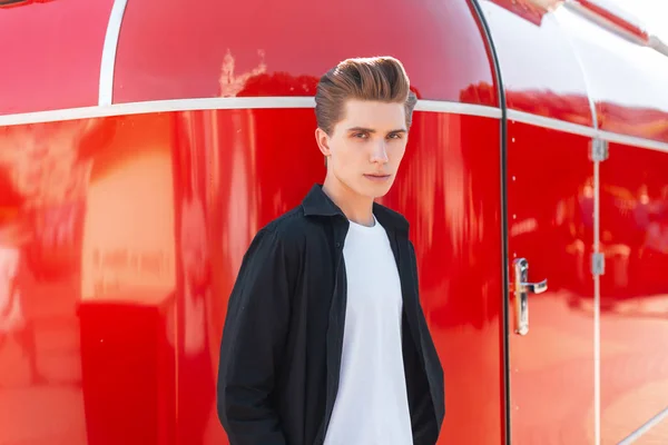 Joven hombre elegante con un peinado de moda en una elegante camiseta con una camisa negra vintage está de pie cerca de una pared de metal rojo en un día soleado de verano brillante. El tipo serio está descansando al aire libre. . — Foto de Stock