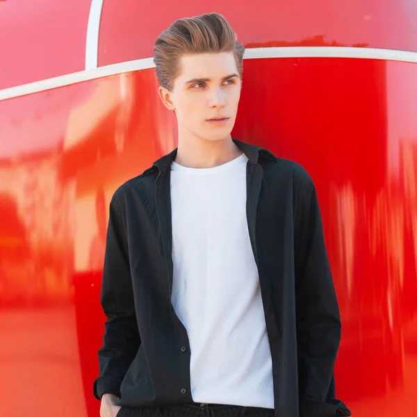 Jeune homme attrayant avec une coiffure élégante dans une chemise noire vintage dans un T-shirt blanc se dresse sur un fond rouge vif dans la ville par une journée d'été ensoleillée. beau mec européen modèle de mode . — Photo