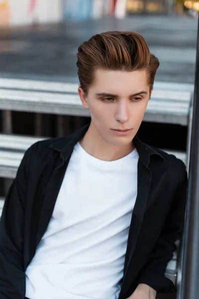 European tânăr atractiv în cămașă neagră elegantă în tricou alb vintage cu coafură la modă așezată pe o scară metalică argintie din oraș. Un tip de treabă se odihneşte într-o zi de vară. Stilul modern — Fotografie, imagine de stoc