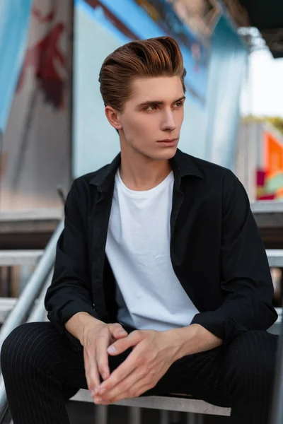 Europese knappe jonge man in een elegant zwart shirt in een wit T-shirt in trendy broek met een stijlvolle kapsel rusten zittend op een metalen trap in de stad. Aantrekkelijk Guy fashion model. — Stockfoto
