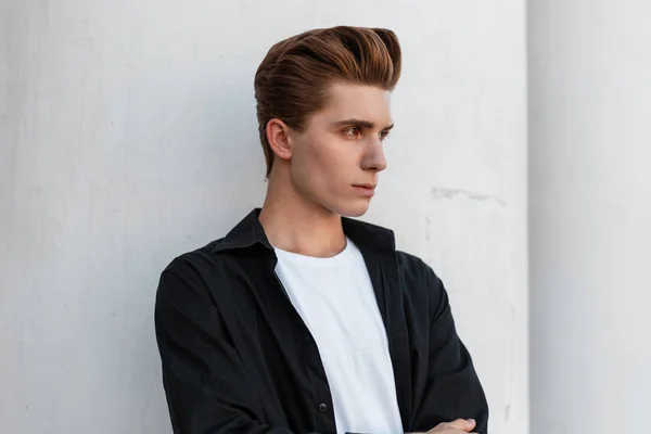 Un joven urbano con un elegante peinado en una elegante camisa negra con una camiseta blanca está de pie cerca de un edificio vintage con columnas blancas en un día de verano. Retrato de hombre de moda al aire libre . — Foto de Stock