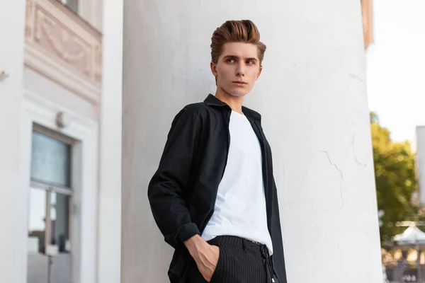 Elegante joven con una camisa negra de moda en una camiseta blanca con estilo en pantalones a rayas con un peinado de moda descansa cerca de una columna blanca vintage. Tipo atractivo en la calle en un día de verano . — Foto de Stock