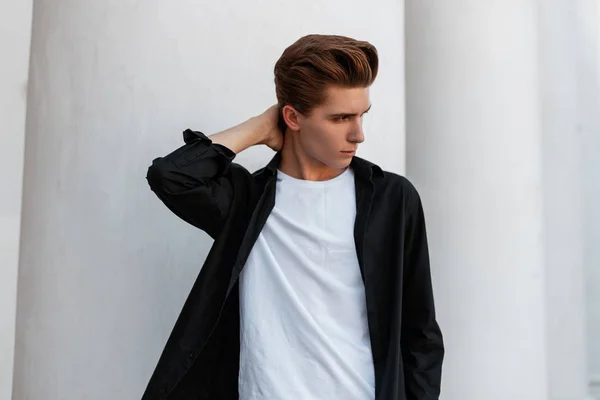 Elegante joven urbano con una camisa negra de moda en una camiseta blanca con un peinado elegante se encuentra cerca de un edificio vintage blanco con columnas en la ciudad. Guapo chico moda modelo al aire libre . — Foto de Stock