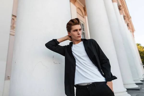 Atractivo joven con un peinado de moda en una camisa negra clásica en una elegante camiseta posando cerca de un edificio con columnas vintage. Guapo chico moderno es relajante al aire libre en la ciudad . — Foto de Stock