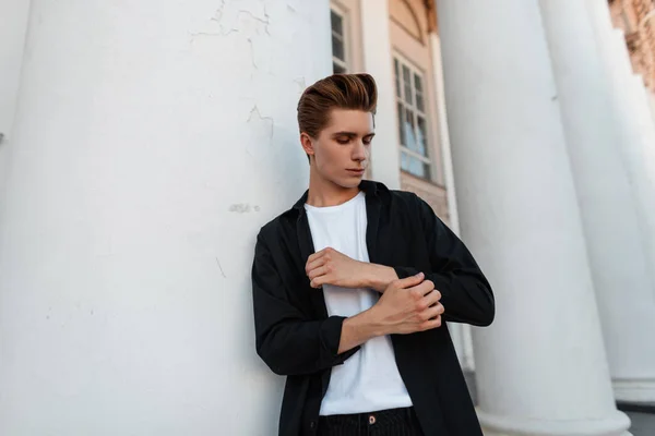 Joven urbano guapo con una elegante camisa negra en una camiseta blanca con un peinado de moda se encuentra cerca de las columnas blancas de un edificio moderno de la ciudad. Atractivo buen tipo descansando en la calle . — Foto de Stock