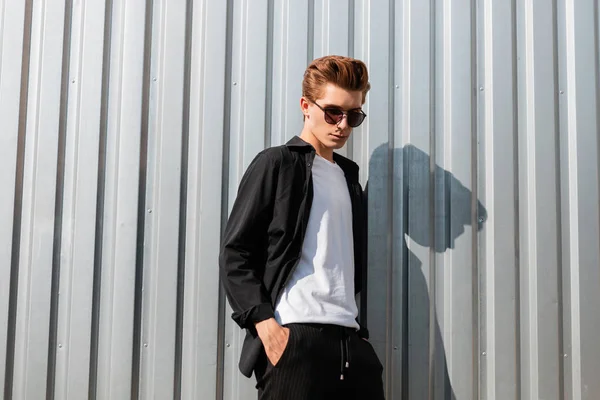 Jonge moderne man hipster in een elegante shirt in een t-shirt in trendy gestreepte broek in zonnebril in de buurt van zilveren metalen wand in de stad. Leuke kerel met een stijlvol kapsel ontspant buiten op een zonnige dag. — Stockfoto