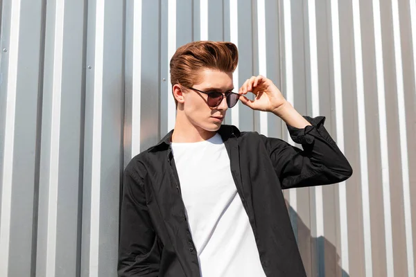 Modelo joven atractivo en una camisa negra de moda en una camiseta endereza gafas de sol con estilo. Un hipster europeo posando cerca de una pared de metal plateado en una ciudad en un día soleado brillante. Estilo moderno — Foto de Stock