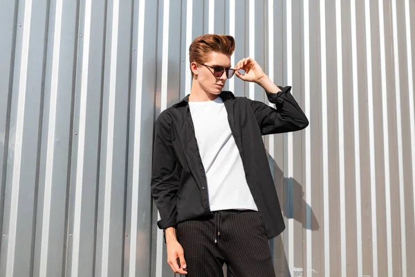 O jovem hipster em calças listradas vintage em uma camisa preta elegante em uma camiseta branca em óculos de sol na moda posa perto de uma parede metálica em um dia ensolarado brilhante. cara urbano moderno gosta do sol de verão . — Fotografia de Stock