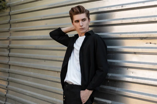 Joven urbano guapo con un peinado de moda en una elegante camisa negra en una camiseta con pantalones a rayas de moda posando cerca de la pared de metal plateado al aire libre. Modelo de moda de hombre moderno. Ropa de hombre con estilo . — Foto de Stock