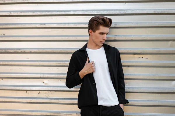 Moderno joven elegante con un peinado elegante en una camisa negra de moda en una camiseta blanca en pantalones posa cerca de una pared vintage de plata en la ciudad. Hombre europeo urbano modelo de moda. Estilo . — Foto de Stock