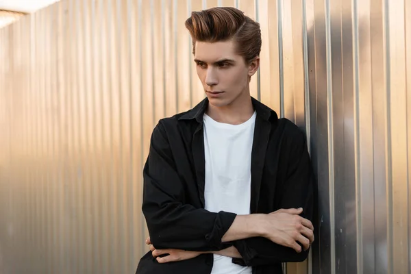 Joven hombre serio con un peinado de moda en una elegante camiseta con una camisa negra vintage está de pie cerca de una pared de metal plateado en un día soleado de verano brillante. Elegante chico modelo está descansando al aire libre . — Foto de Stock