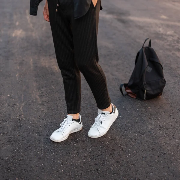 Primer plano de las piernas masculinas. Joven hombre de moda en pantalones negros vintage en cuero elegantes zapatillas blancas posando de pie sobre el pavimento contra una mochila negra. Estilo de calle juvenil moderno . —  Fotos de Stock