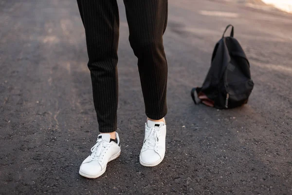 Jovem elegante fica no pavimento em couro tênis branco em calças pretas listradas vintage calças com uma mochila preta na moda. Roupa de homem sazonal na moda e sapatos. Close-up . — Fotografia de Stock